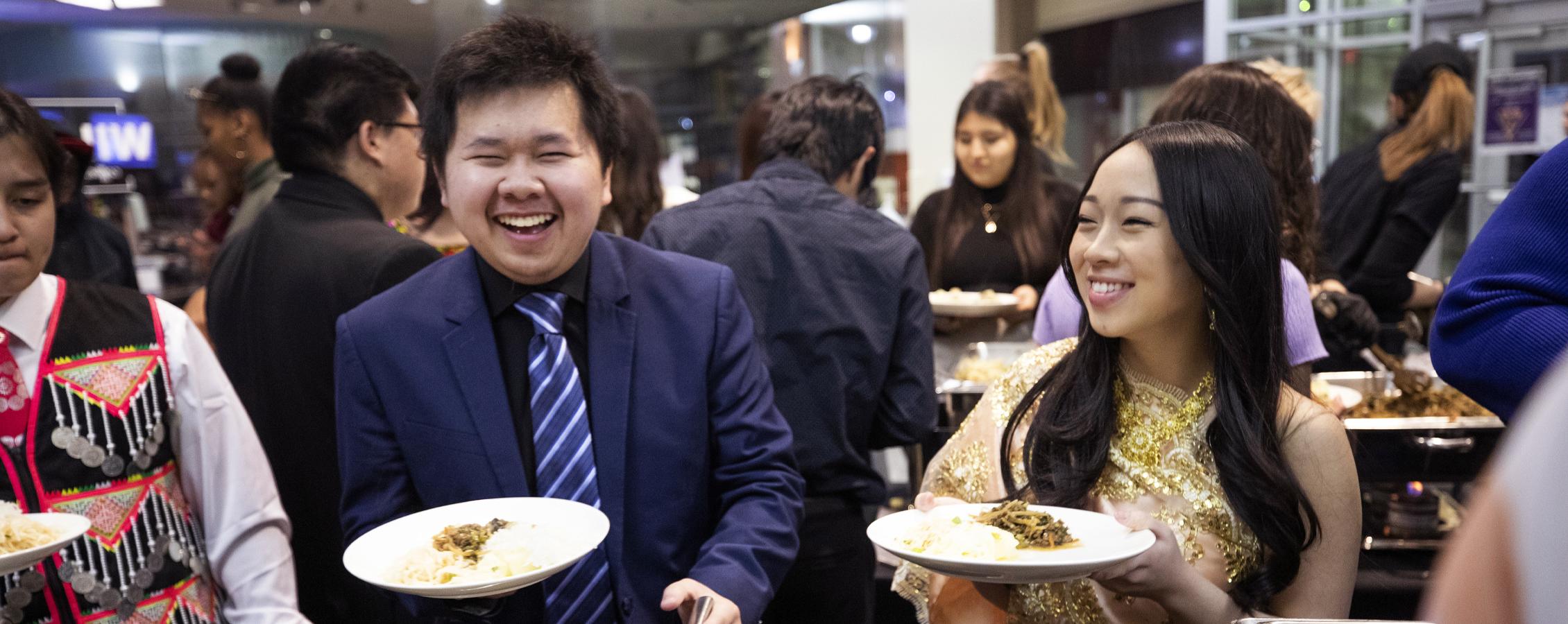 Two students smile with plates of food in their hands.