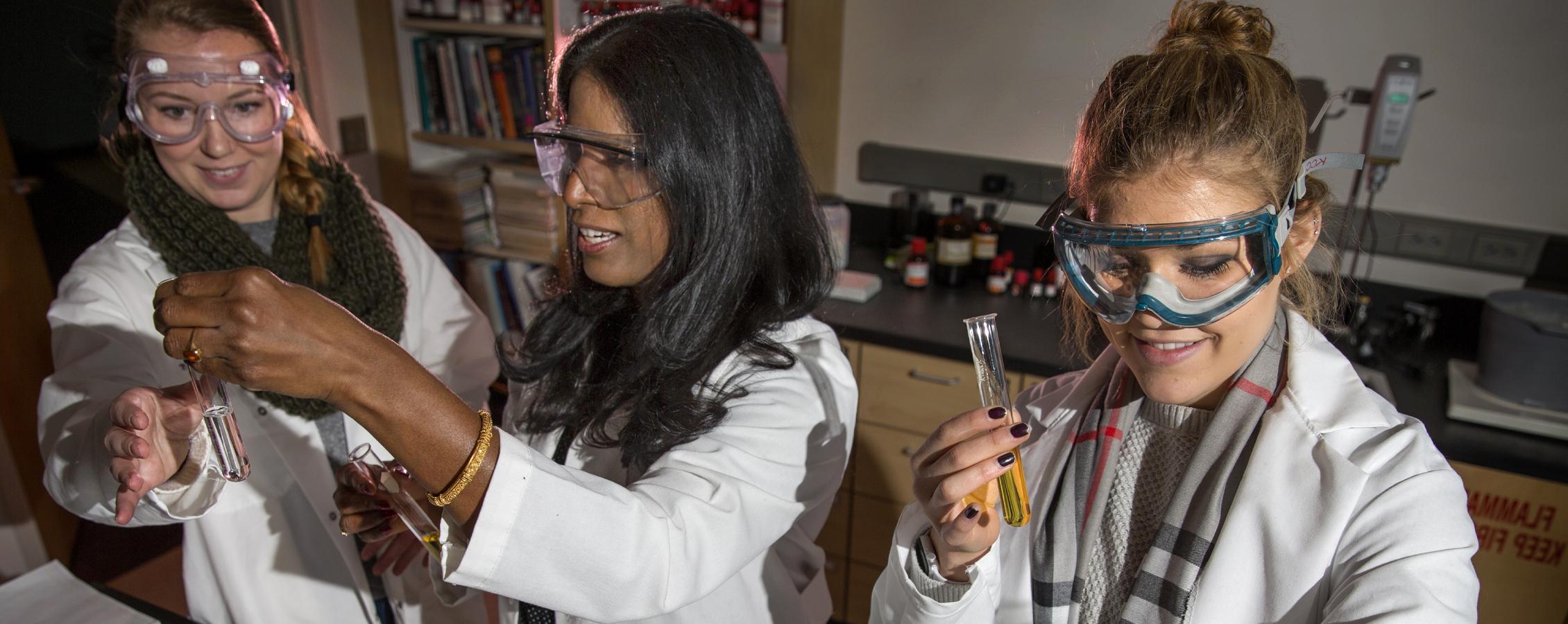 A faculty member and two students, all in lab coats, look at liquid vials.