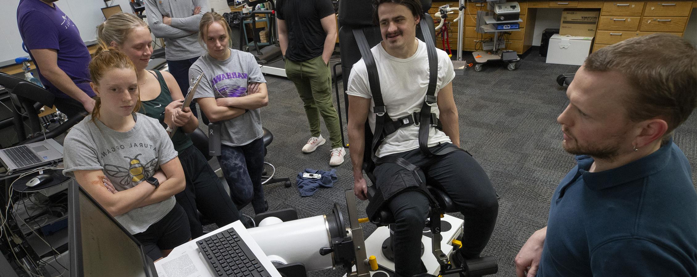 A faculty member and students gather around a student strapped in a chair to collect data during an exercise during class.